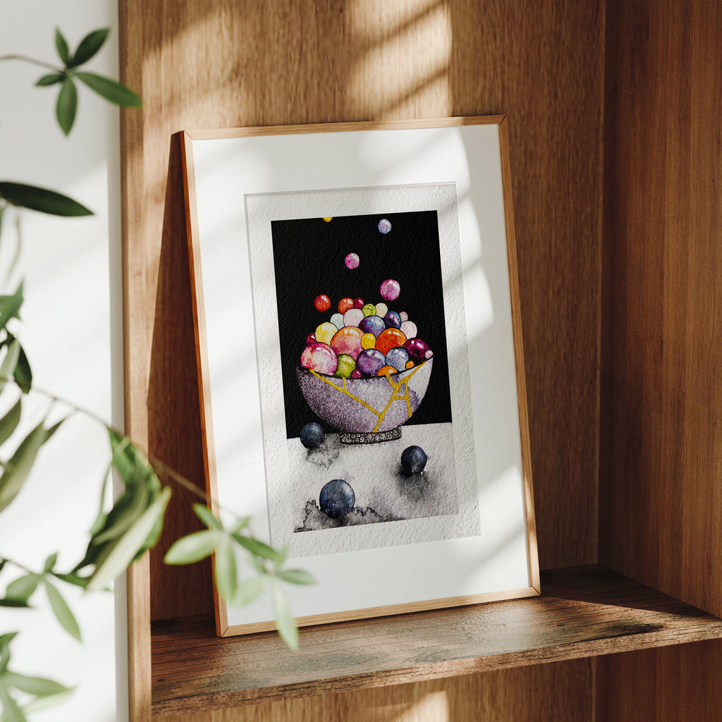 Framed Kintsugi fine art print on a wooden shelf in dappled daylight, a gold-repaired grey bowl overflowing with glossy coloured spheres against deep black, by Léna Ruisz.