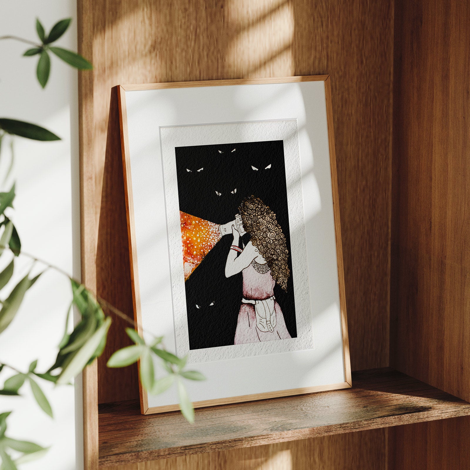 Framed Making Light fine art print on a wooden shelf in dappled daylight, a woman with a camera casting a cone of golden light into darkness ringed by glowing eyes, by Léna Ruisz.