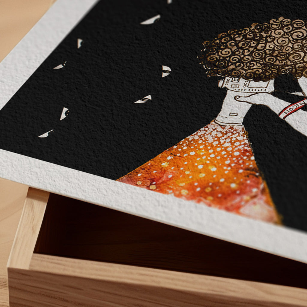 Detail of the Making Light fine art print resting on a wood surface, showing the deckle paper edge and close-up of a curly-haired figure casting a stream of golden light against a black ground in ink and watercolour