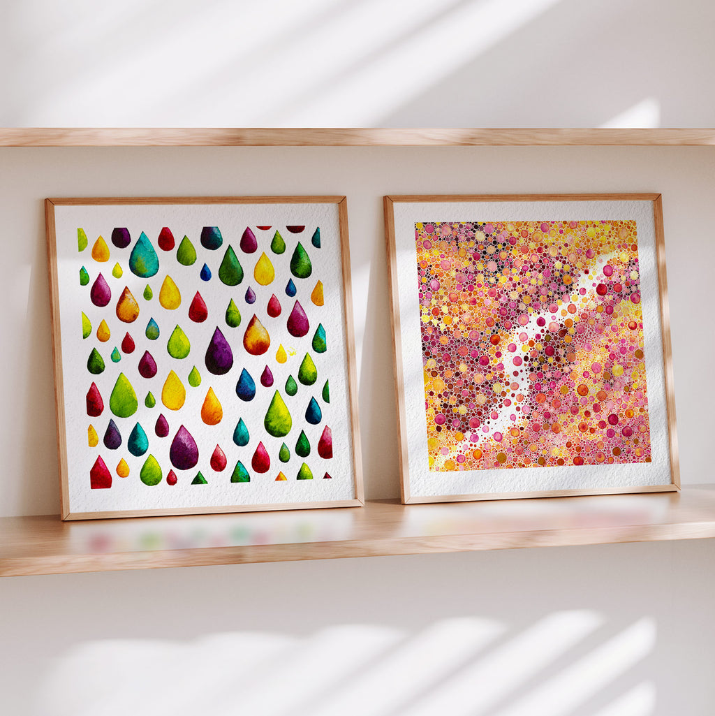 Two framed Materia fine art prints on a wooden shelf in soft daylight: Flow, scattered rainbow watercolour raindrops on white, beside Prima Materia, a dense field of pink, gold and magenta watercolour dots threaded by bare paper.