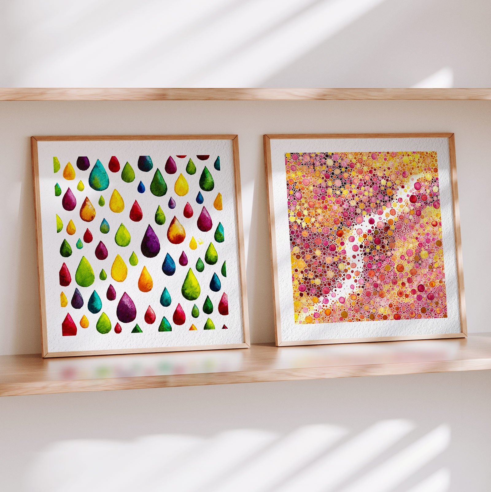 Two framed Materia fine art prints on a wooden shelf in soft daylight: Flow, scattered rainbow watercolour raindrops on white, beside Prima Materia, a dense field of pink, gold and magenta watercolour dots threaded by bare paper.