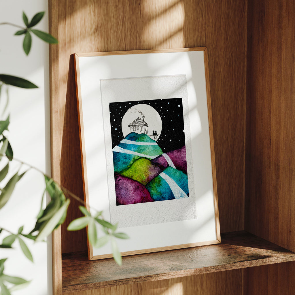 Framed Our Life Pictured fine art print on a wooden shelf in dappled daylight, a tiny couple seated by a moonlit hilltop house above layered patchwork hills, by Léna Ruisz.