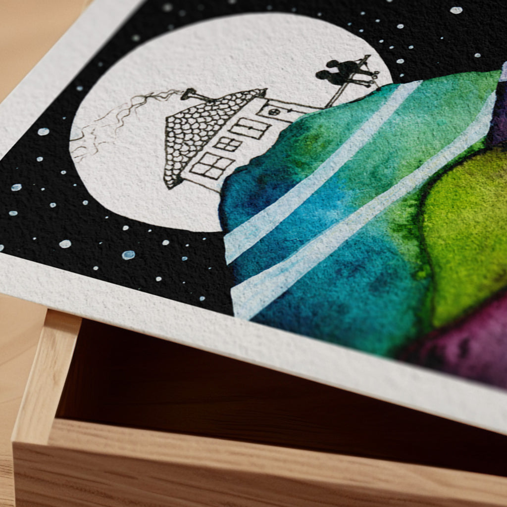 Detail of the Our Life Pictured fine art print resting on a wood surface, showing the deckle paper edge and close-up of a small house beneath a full moon above hills of teal and green watercolour in ink and watercolour
