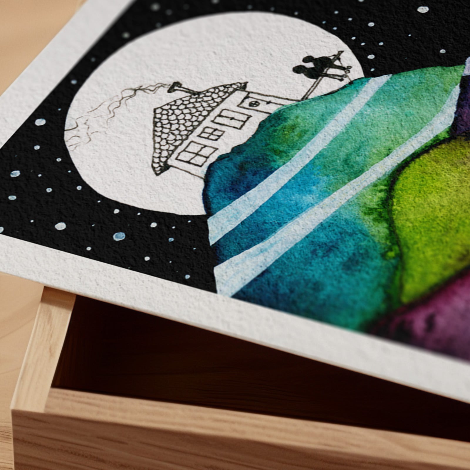 Detail of the Our Life Pictured fine art print resting on a wood surface, showing the deckle paper edge and close-up of a small house beneath a full moon above hills of teal and green watercolour in ink and watercolour
