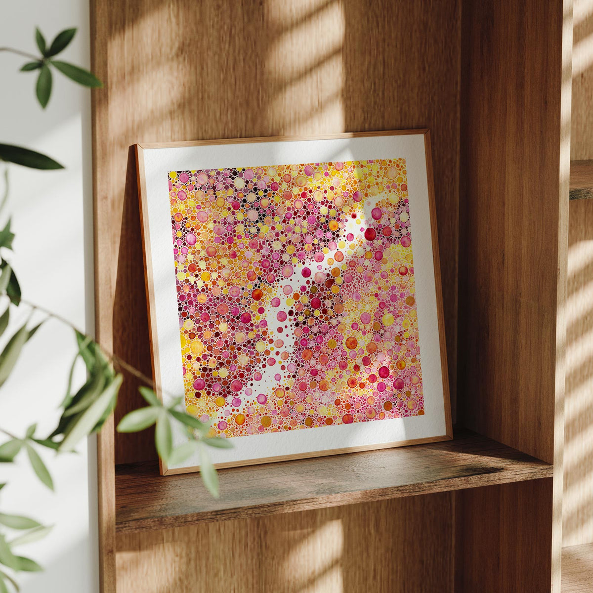 Framed Prima Materia fine art print leaning on a wooden shelf in dappled daylight, a dense field of pink, gold and magenta watercolour dots parted by a winding channel of bare paper, by Léna Ruisz.
