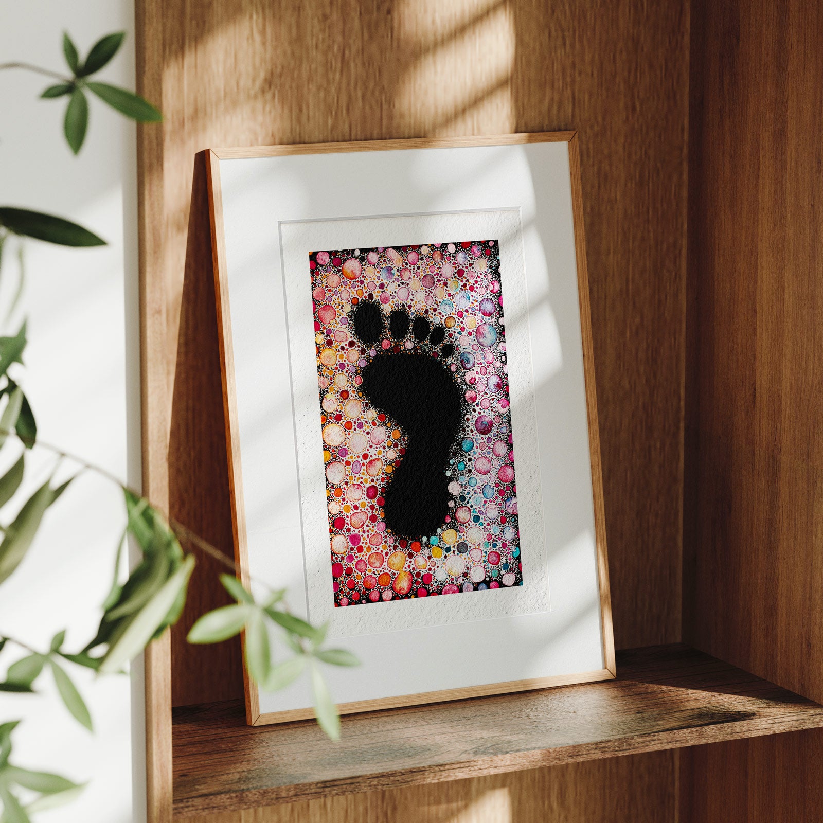 Framed Spoor fine art print on a wooden shelf in dappled daylight, a footprint formed as dark negative space within a dense field of colourful watercolour dots, by Léna Ruisz.