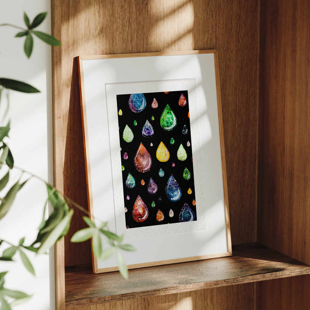 Framed Still Falling fine art print on a wooden shelf in dappled daylight, translucent crackle-glazed raindrops in many colours falling through deep black, by Léna Ruisz.
