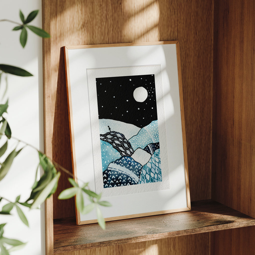 Framed The Watcher fine art print on a wooden shelf in dappled daylight, a lone figure on a rocky outcrop beneath a full moon and a star-scattered night sky, by Léna Ruisz.